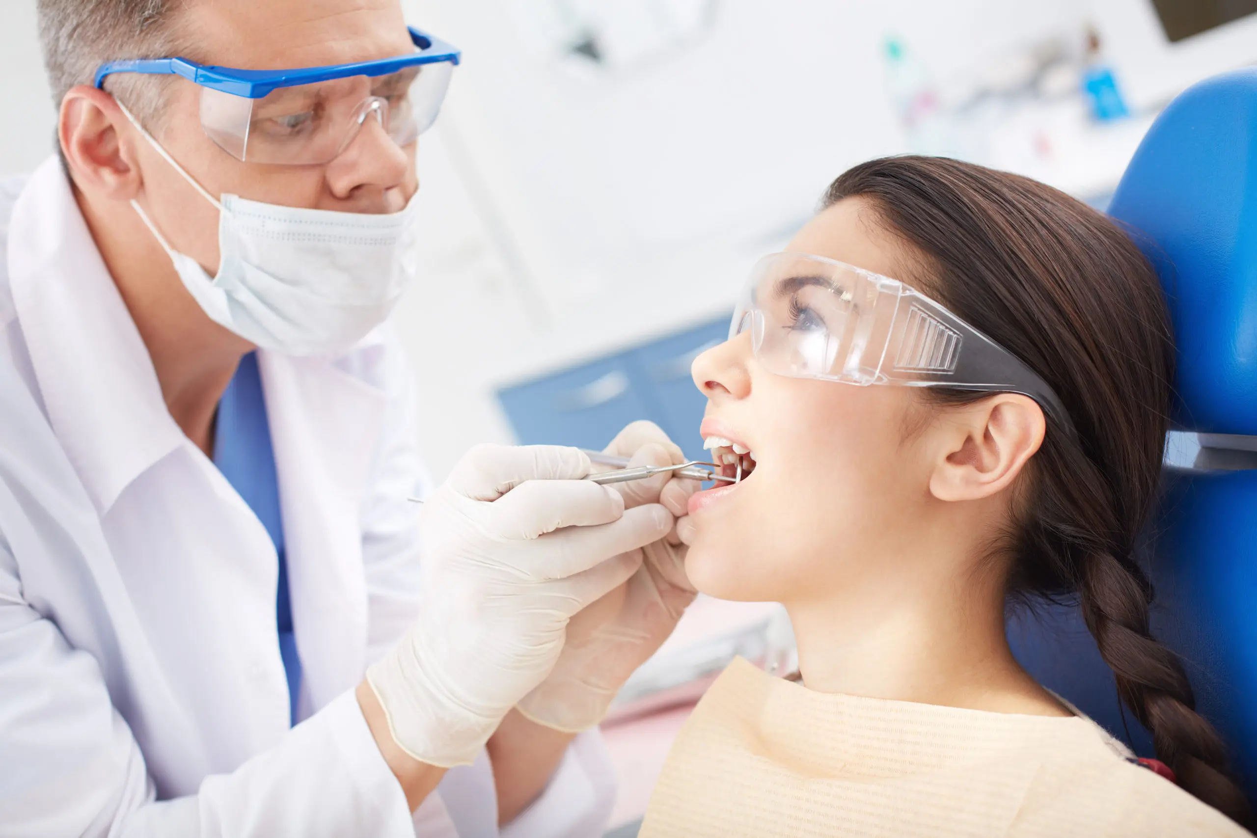 A dentist in protective eyewear and a mask examines a patient's teeth with tools. The patient, wearing protective glasses, sits in a dental chair.