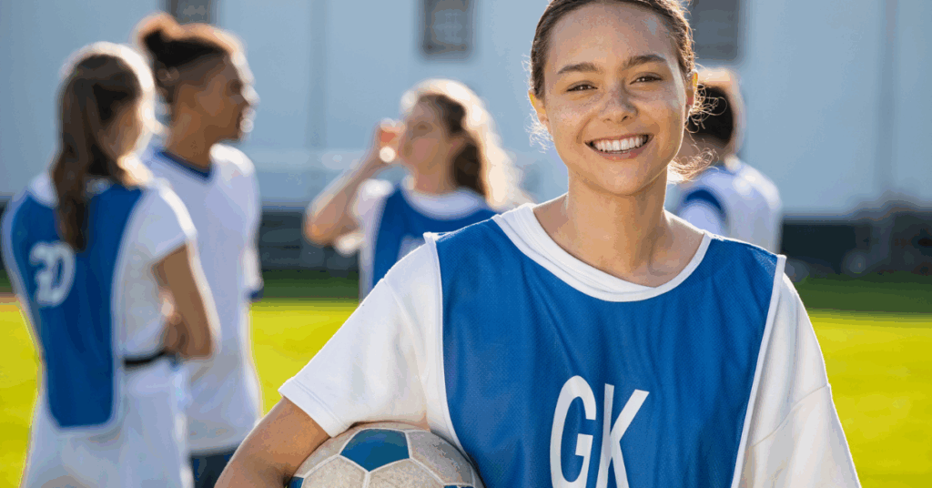 A girl in a blue uniform stands holding a soccer ball, ready for play on a grassy field.