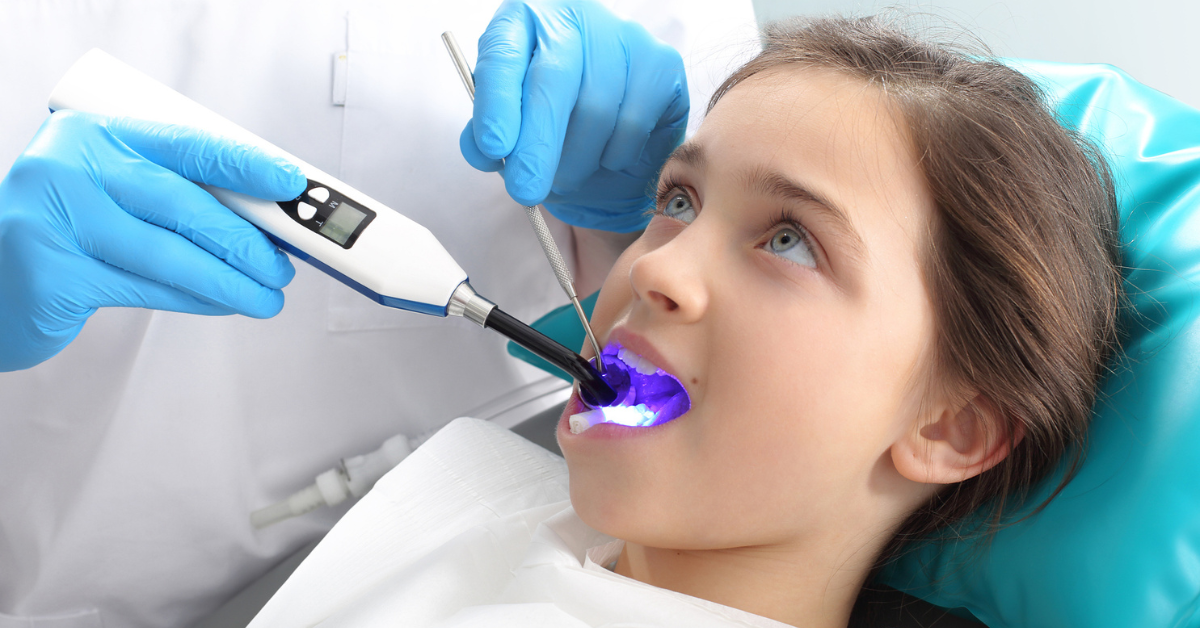 A dentist in blue gloves uses a curing light inside a young girl's mouth during a dental sealent procedure. The girl, lying back, looks calm and curious.