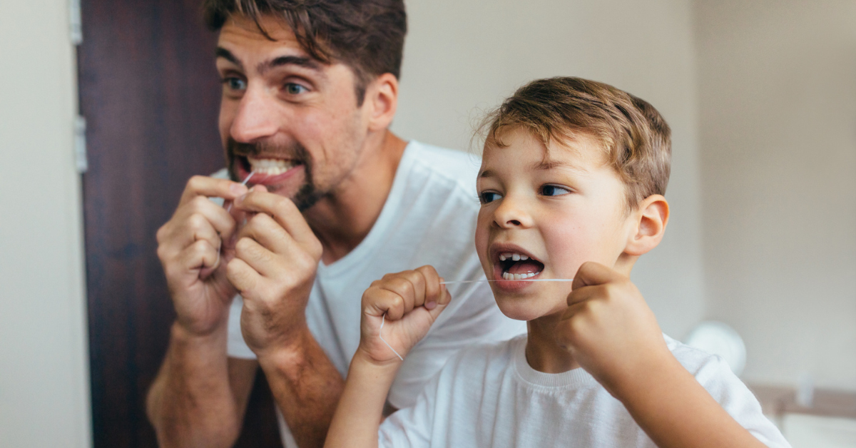 Father and son in white shirts flossing their teeth together in a bathroom, smiling in a playful and engaging moment of dental care routine.