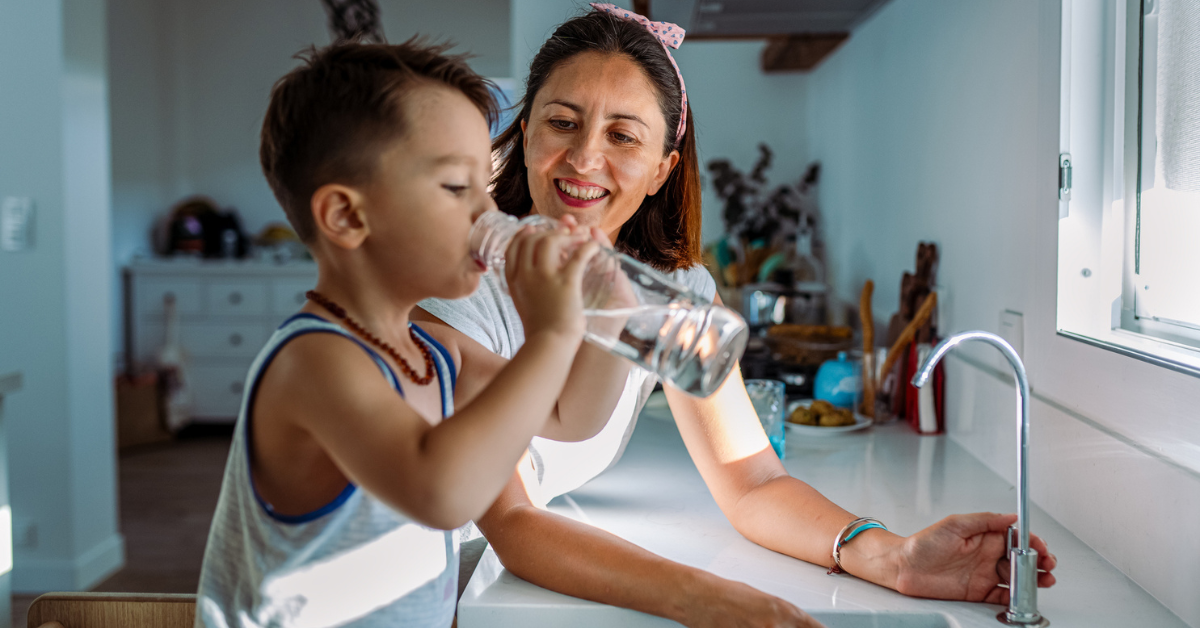 A mother smiles warmly at her young son as he drinks water from a glass bottle in a bright kitchen. The atmosphere is joyful and nurturing.