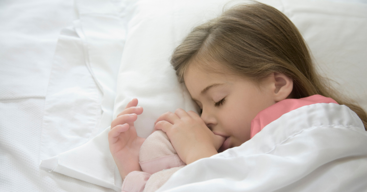 A young girl sleeps peacefully in bed, sucking her thumb and cuddling a pink stuffed animal. She wears a white blanket and pink pajamas, conveying comfort and serenity.