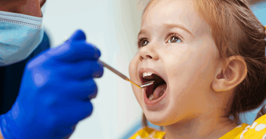 A dentist, wearing a mask and gloves, examines a young girl’s open mouth with a dental mirror. The girl, in a yellow top, appears curious and calm.