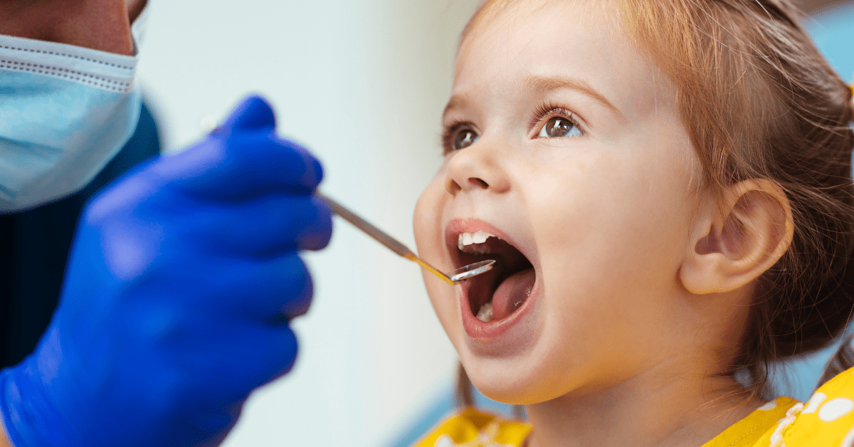 A dentist, wearing a mask and gloves, examines a young girl’s open mouth with a dental mirror. The girl, in a yellow top, appears curious and calm.