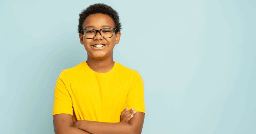 Smiling boy with glasses and a bright yellow shirt stands confidently against a pale blue background, conveying a cheerful and vibrant mood.