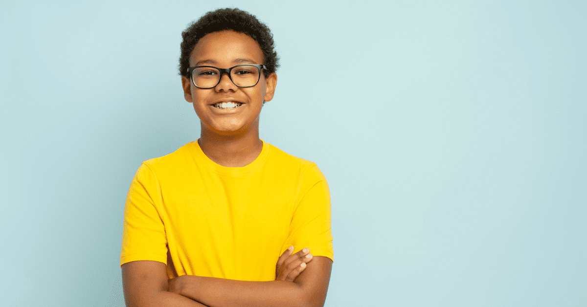 Smiling boy with glasses and a bright yellow shirt stands confidently against a pale blue background, conveying a cheerful and vibrant mood.