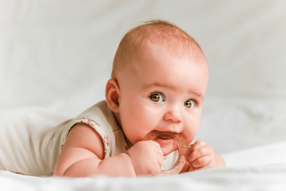 A baby lying on a white blanket is chewing on a teething ring. The infant has wide eyes and a calm expression, showcasing an innocent curiosity.