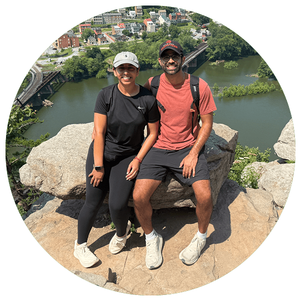 A smiling couple sits on a rock ledge overlooking a scenic river and town. They're dressed in casual hiking attire, exuding a cheerful, relaxed vibe.