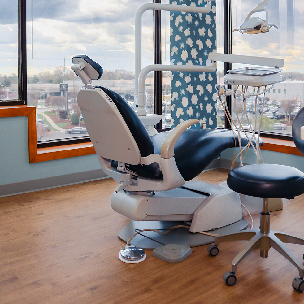 Dental office with a modern chair, blue upholstery, and nearby equipment against a large window. Calm ambiance with wooden flooring and patterned wallpaper.