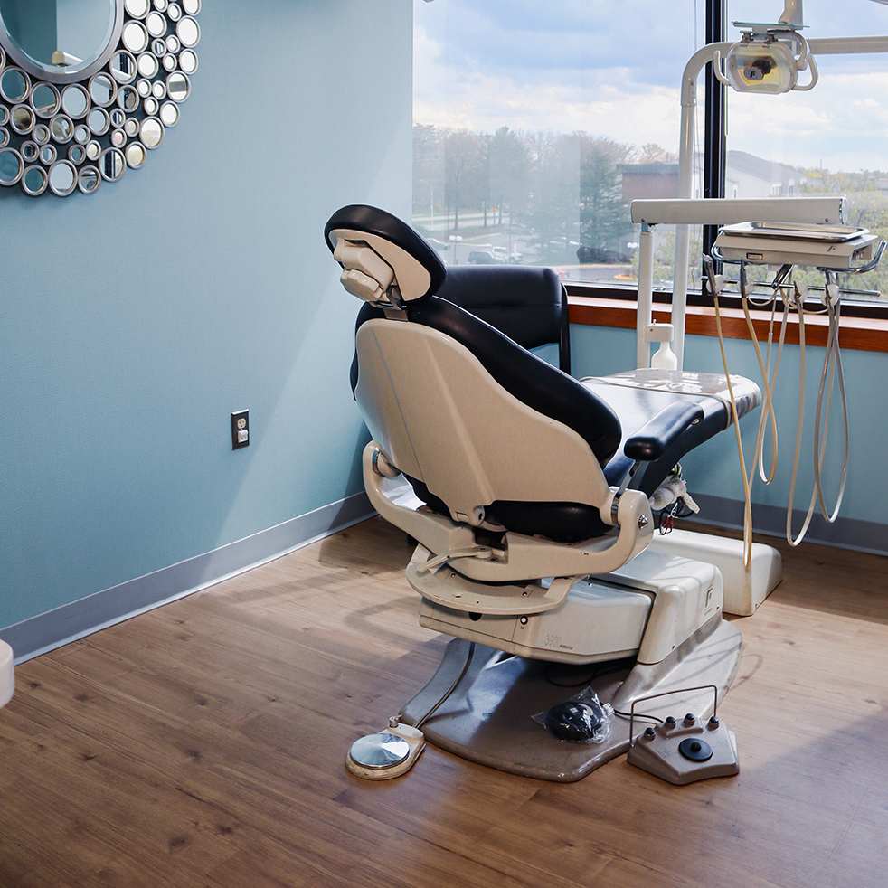 Dental office with a large window view, featuring a modern dental chair in beige and black leather, bright walls, and wooden floors conveying cleanliness.