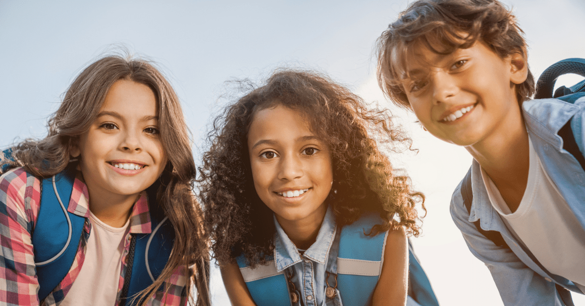 Kids Dentist - Three smiling children with backpacks look into the camera against a clear sky. Their expressions convey happiness and friendship.