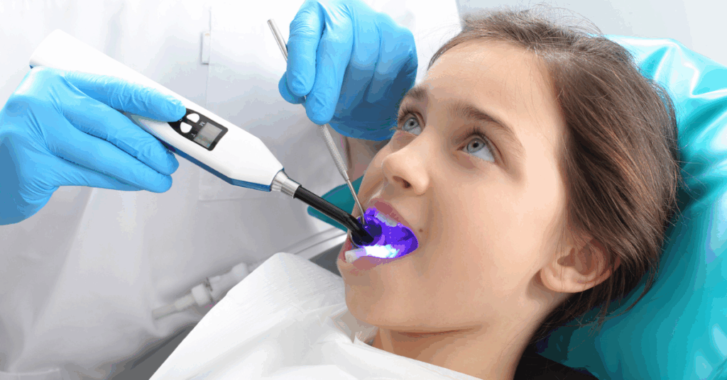 A dentist in blue gloves uses a curing light inside a young girl's mouth during a dental sealent procedure. The girl, lying back, looks calm and curious.