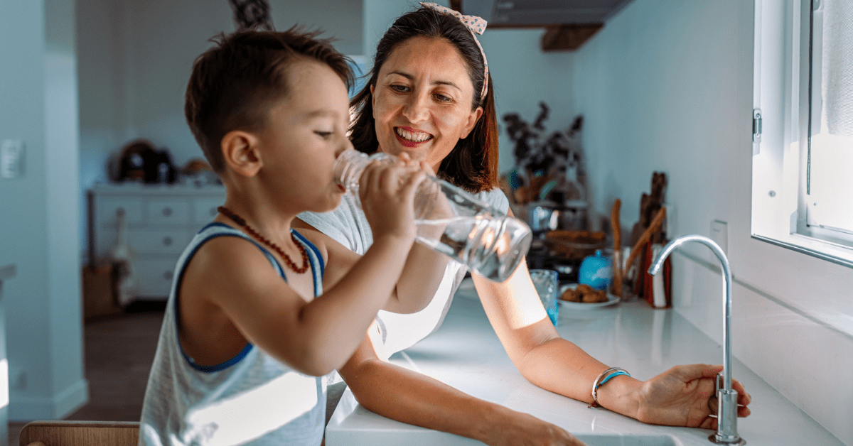 A mother smiles warmly at her young son as he drinks water from a glass bottle in a bright kitchen. The atmosphere is joyful and nurturing.