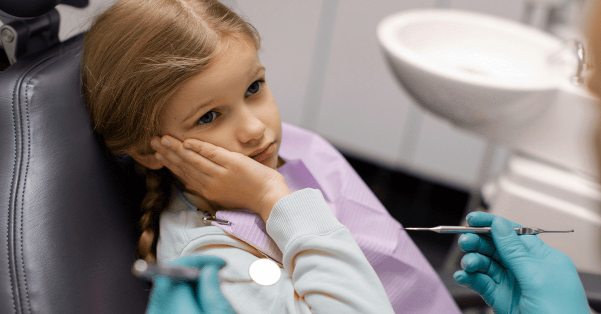 A young girl with a concerned expression sits in a dentist's chair, resting her cheek on her hand. A dentist in gloves holds tools nearby.