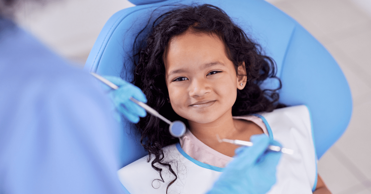 A young girl smiles while sitting in a blue pediatric dental chair during a check-up. A dentist in blue gloves holds dental tools, creating a calm atmosphere.